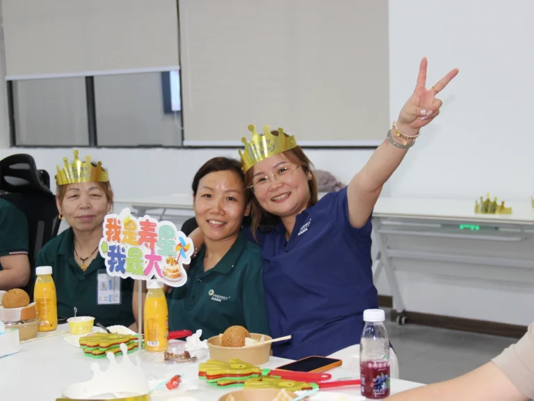 Close-up of smiling birthday employees at office birthday party with golden crowns, birthday table sign, snacks and drinks on table