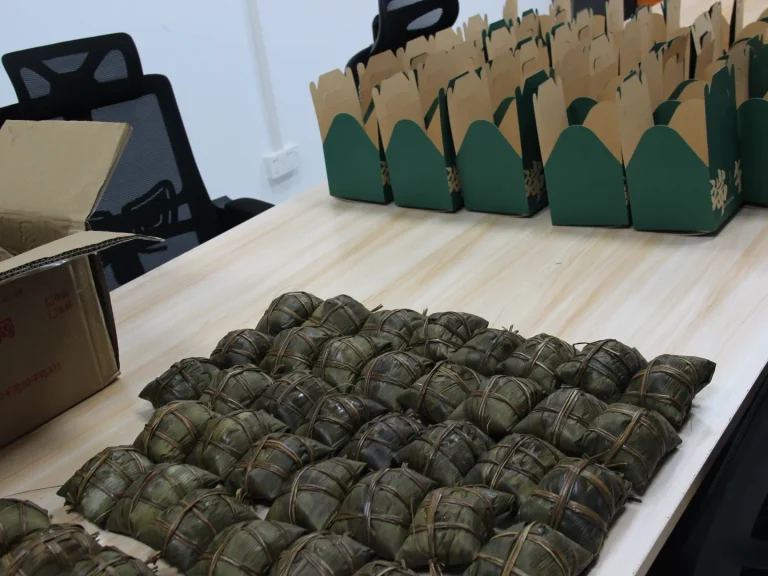 Office preparation shot for Patmat Dragon Boat Festival benefits, piles of handmade rice dumplings matched with green kraft festival gift boxes
